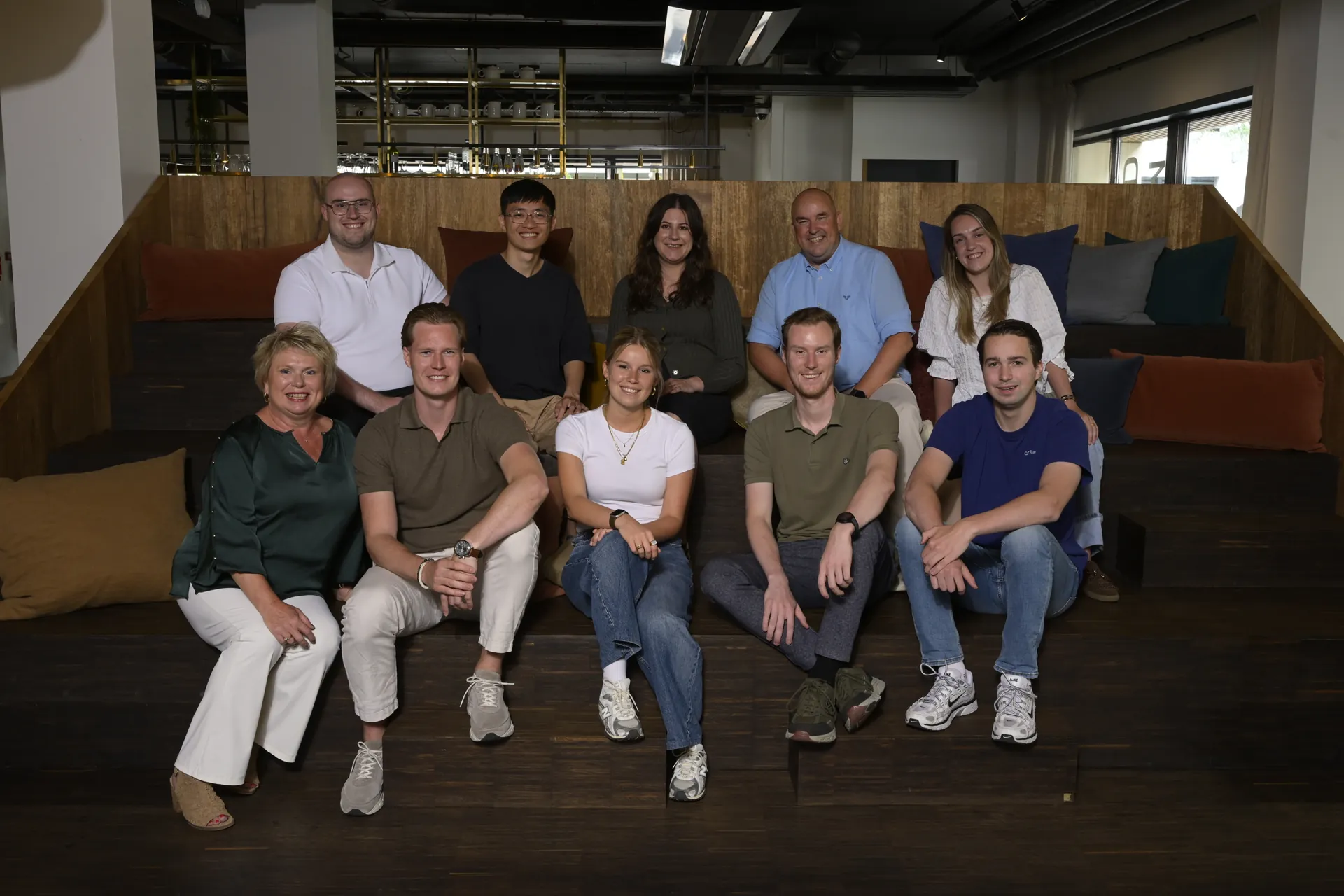 Het AIMAZE-team zit lachend op een brede houten trap in een moderne kantoorruimte.