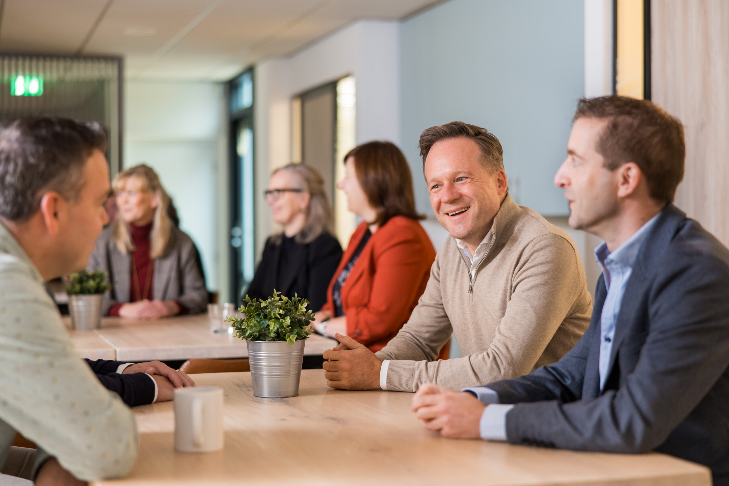 Lachende medewerkers van Dinkelberg & Partners zitten samen aan een vergadertafel in een kantoor, gefotografeerd door een glazen wand.