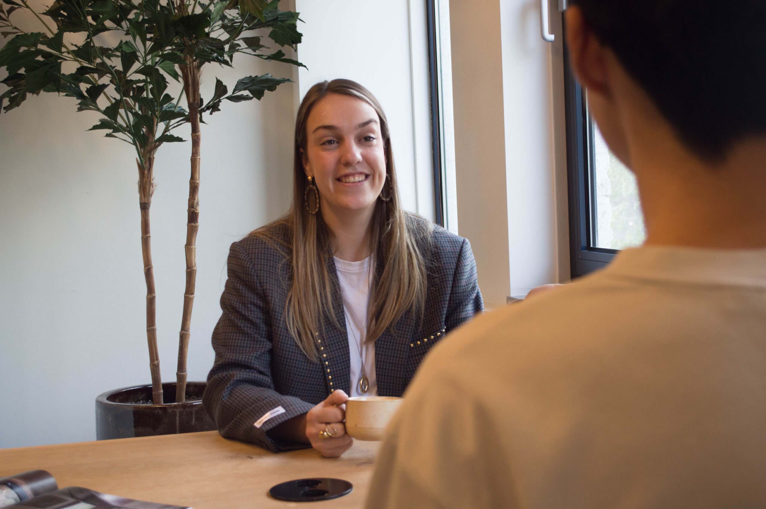 Lachende vrouw zit met een mok aan een houten tafel in een kantoor, met een grote kamerplant bij het raam en een onscherpe gesprekspartner op de voorgrond.